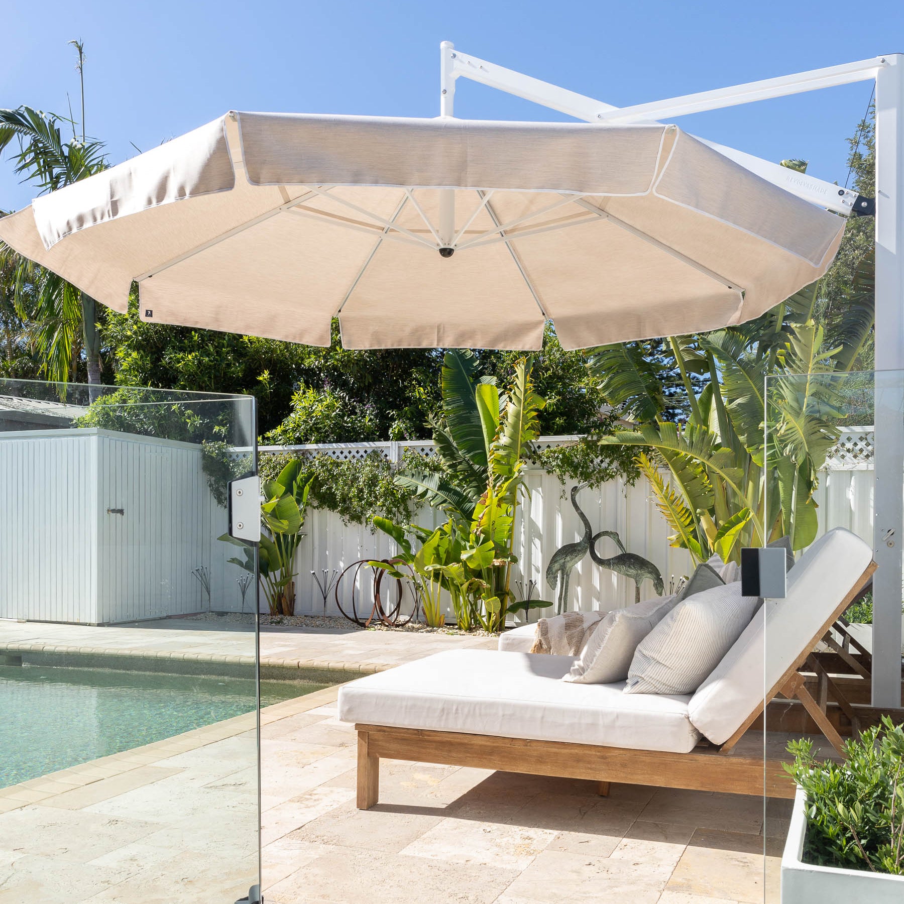 Beige patio umbrella over a lounge chair by a pool with a garden view.